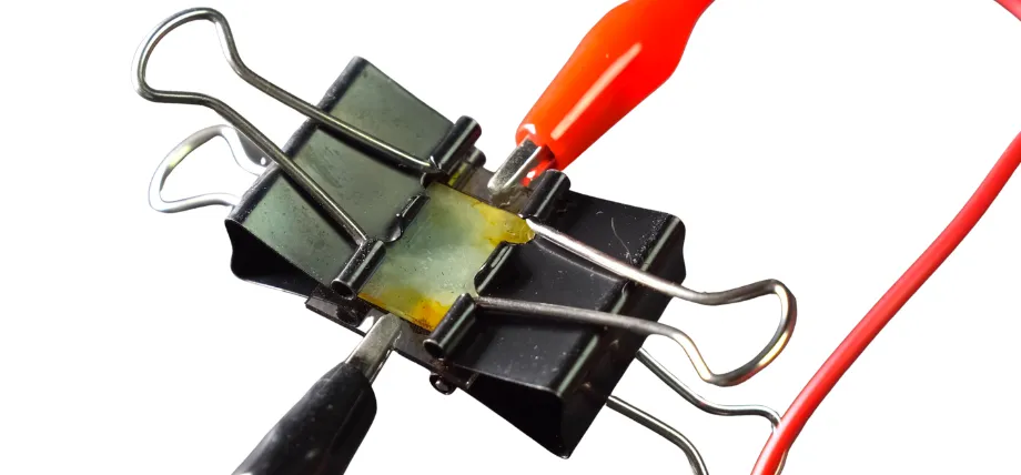 Close-up of a small component held by two black binder clips with red and black alligator clips attached for electrical testing.