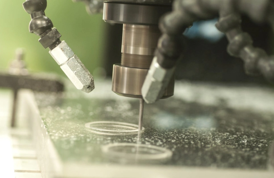 An image of a CNC milling machine cutting circular holes on a translucent sheet with two coolant nozzles directed on the spot 