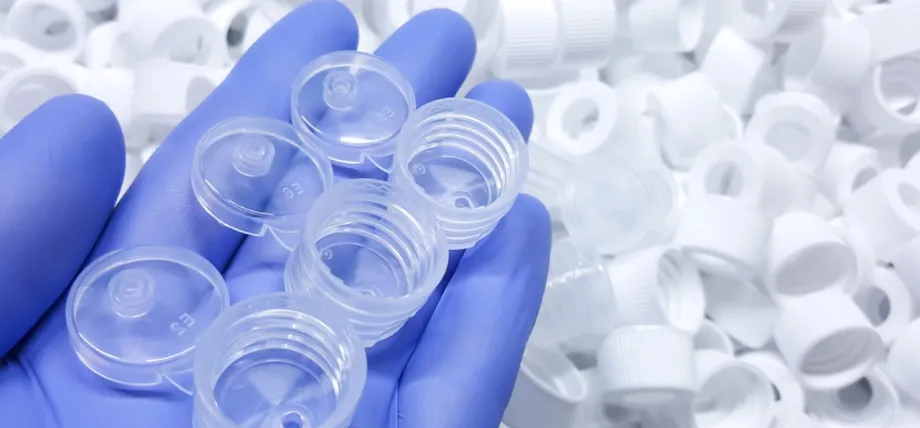 A person wearing blue nitrile gloves holds several clear, threaded plastic medical caps over a pile of white caps.
