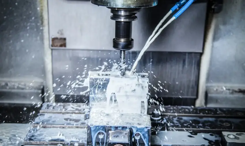 An image of two blue nozzles spraying white substance called coolant on a metal block being CNC drilled