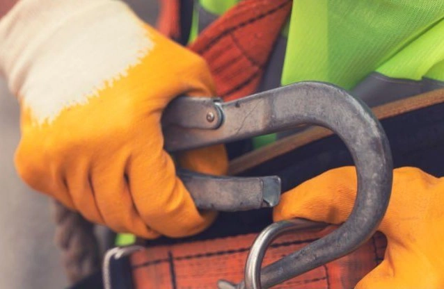 A hand in yellow/white gloves holding a grey carabiner and hooking it to a harness on the waist