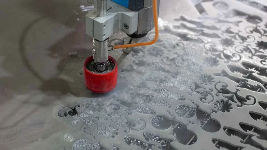 The image shows a precision waterjet cutting machine creating intricate designs on a material. The focused high-pressure water stream cuts through the material, forming detailed and complex patterns.