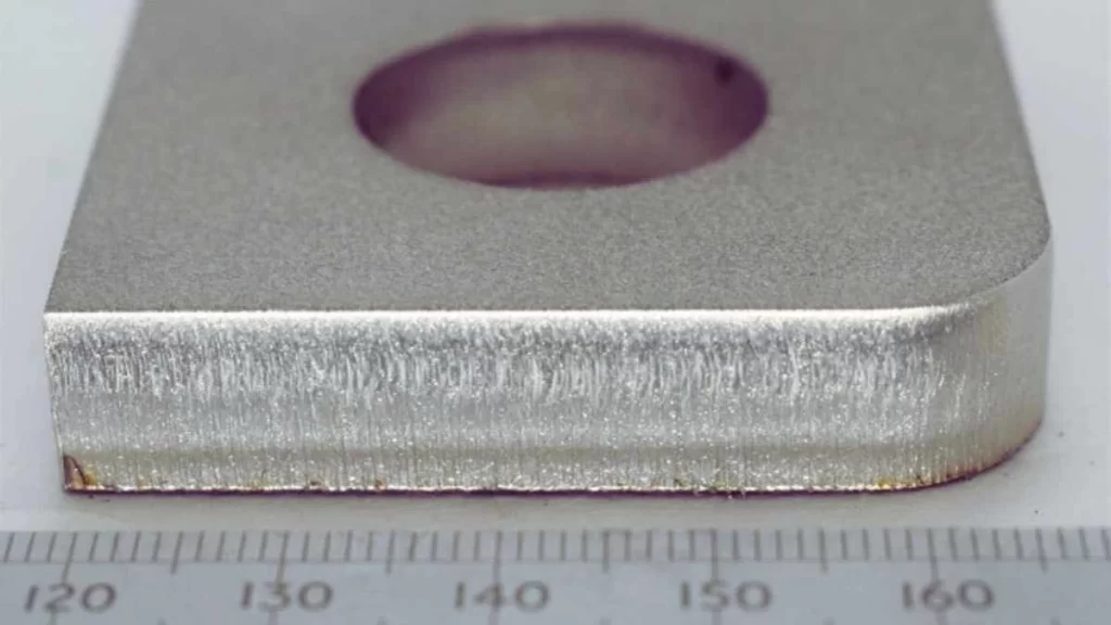 A close-up of a stainless steel piece being cut with a laser. It showcases precise cuts, and a ruler/measuring scale is visible next to the steel, indicating the measurement.