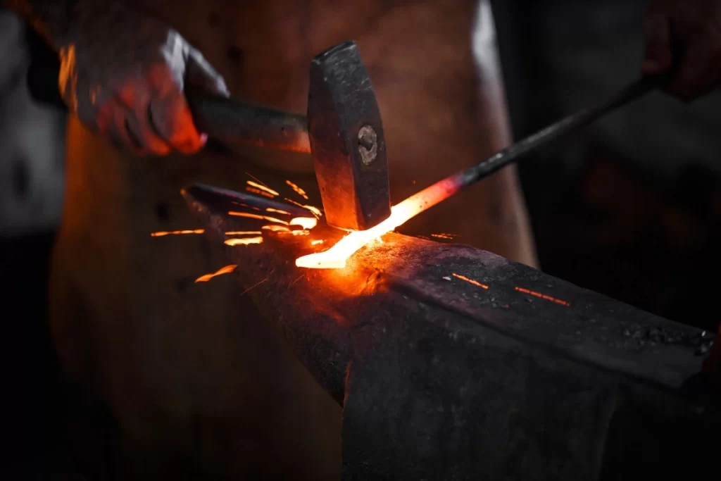 A closeup image showing a man hitting a red hot metal placed on an anvil