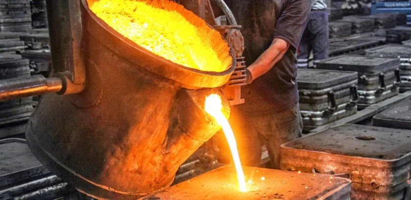 A person pouring hot molten metal from a large metal container into a hole in a material held in a metalling box