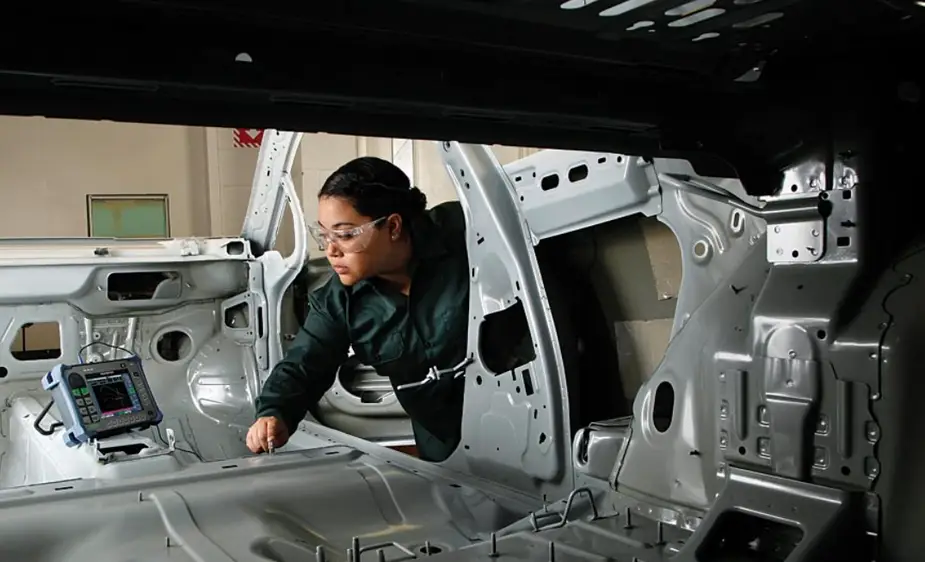 A person in a green overall running diagnostics on the floor of an unfinished car