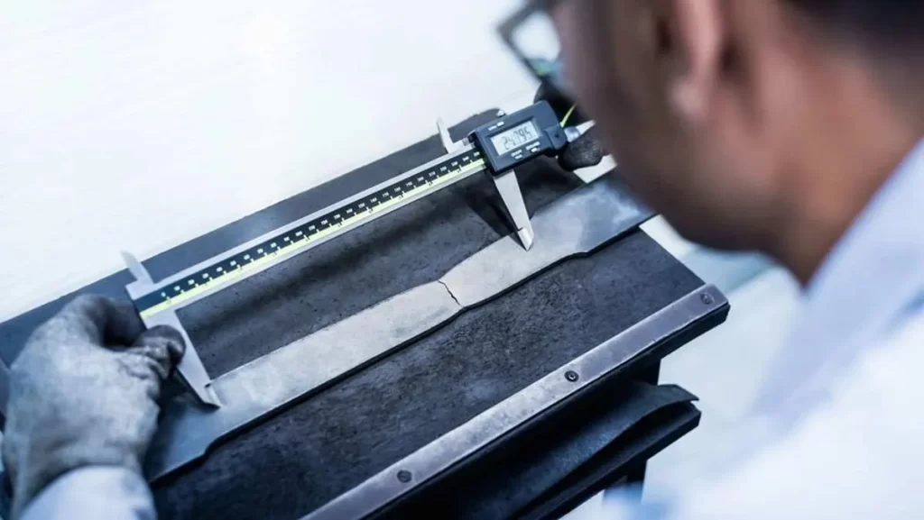 A person in a white lab coat and gloves measuring a fractured metal specimen.