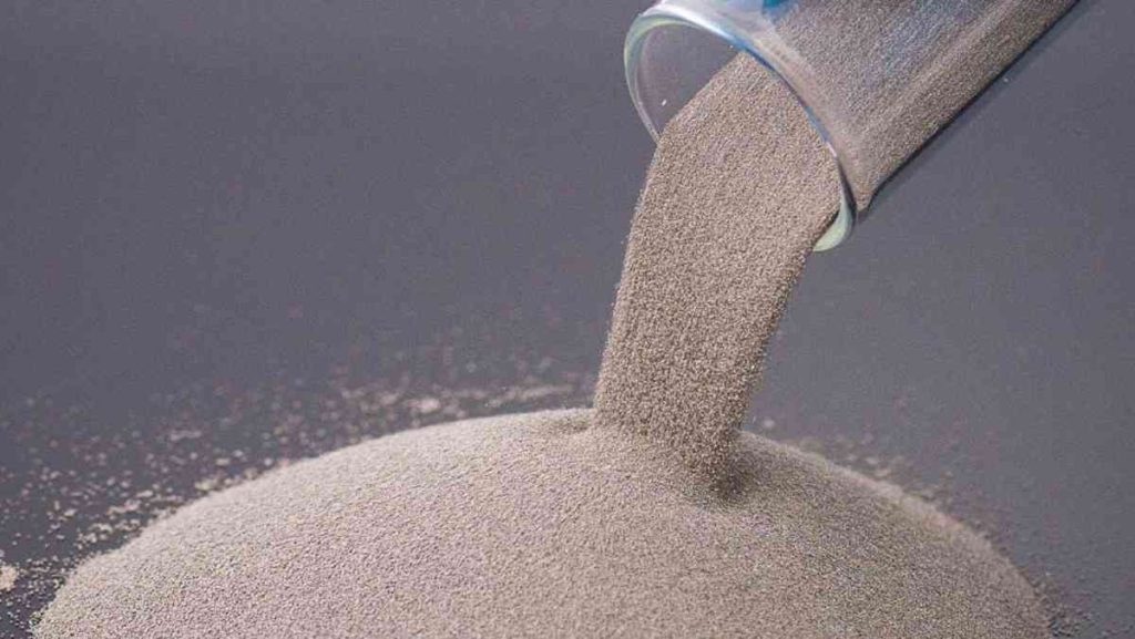 Grey metal powder pouring out of a glass beaker onto a pile