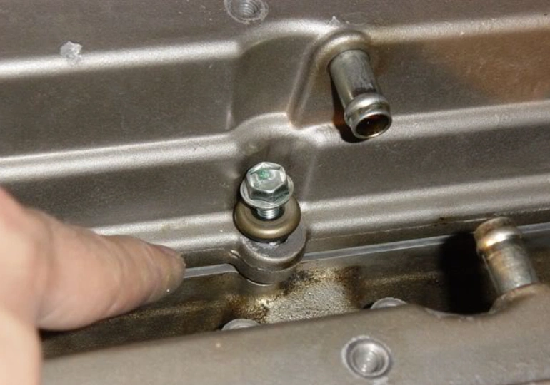 Close-up image showing a fastener with a washer securing an engine valve cover, with a finger pointing at the washer