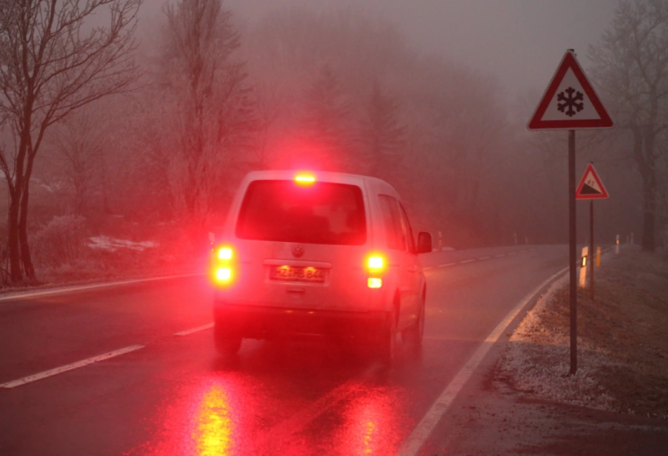 White SUV driving on a wet foggy road with red brake lights illuminated and triangular warning signs visible on the roadside