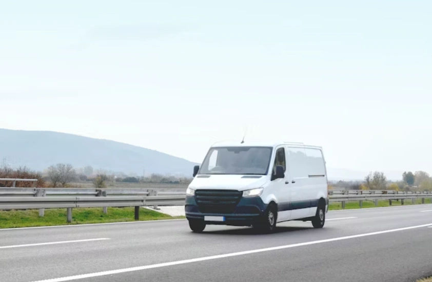 White delivery van with daytime running lights on driving on a highway with mountains and a cloudy sky in the background