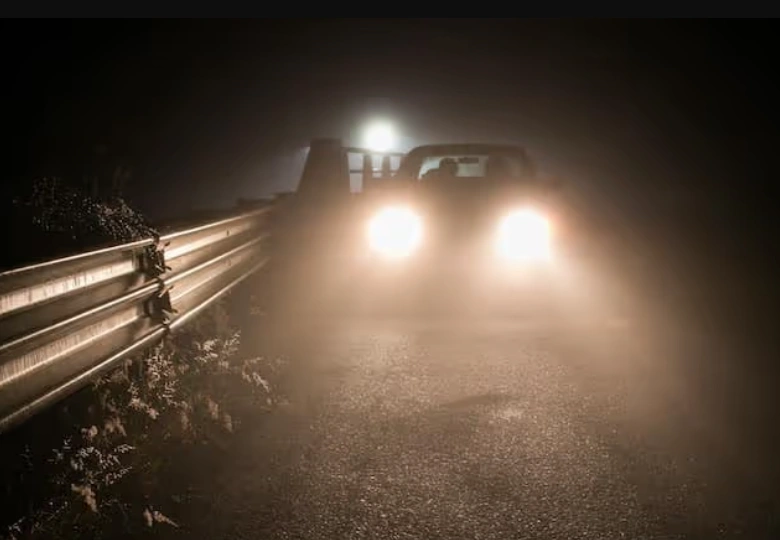 A car driving at night with bright headlights illuminated on a road with metal guardrails 