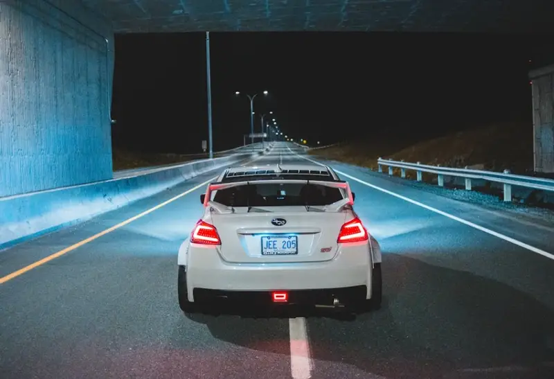 White Subaru driving through a tunnel at night with red tail lights illuminated and street lights visible ahead  