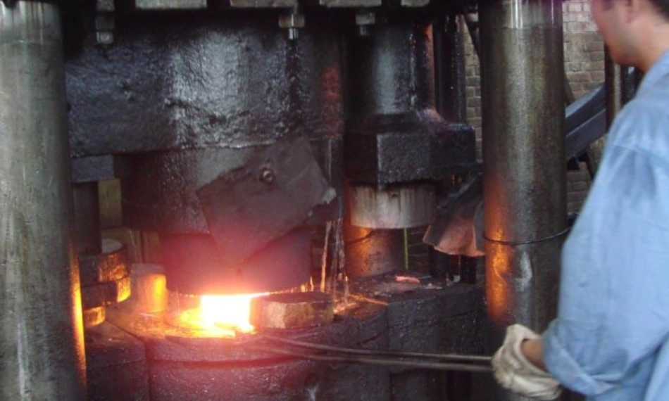 Worker operating a hot forging press with glowing heated metal being shaped under the hammer