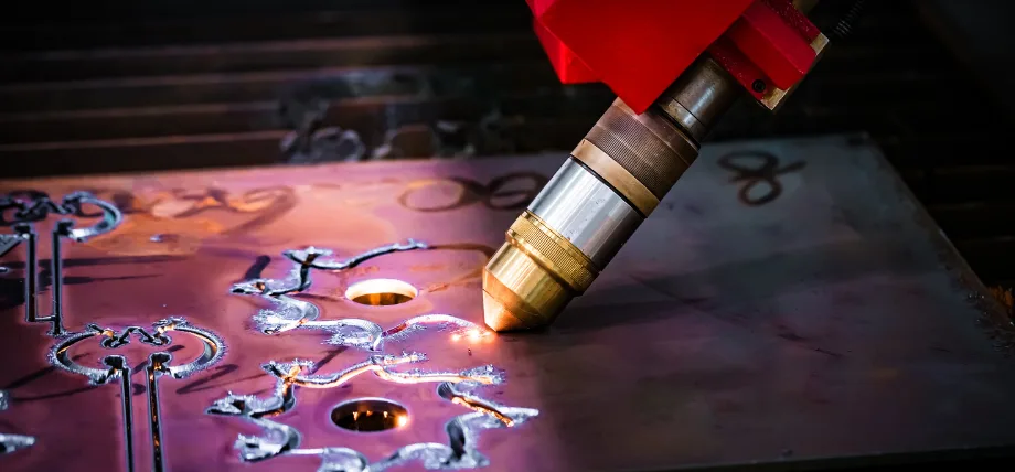 Fiber laser head actively cutting intricate patterns into reflective metal sheet with bright molten glow.