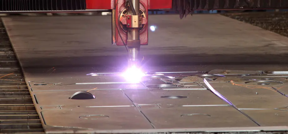 Plasma cutting torch emitting bright white-purple arc and sparks while precisely cutting complex patterns through thick steel plate.