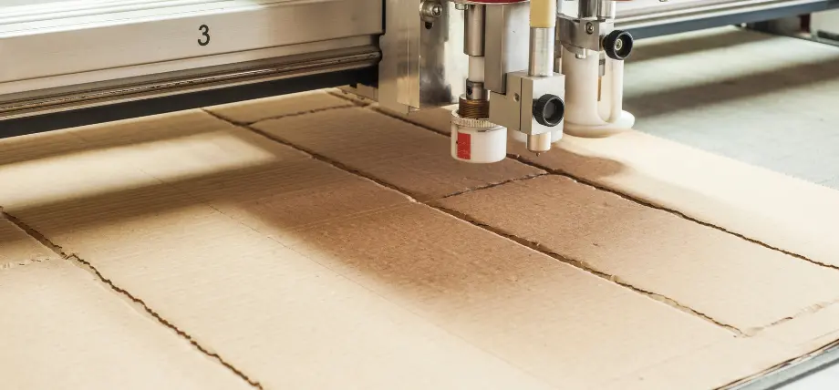 Automated cutting plotter with drag knife tool scoring and cutting corrugated cardboard into clean sections on a vacuum table.