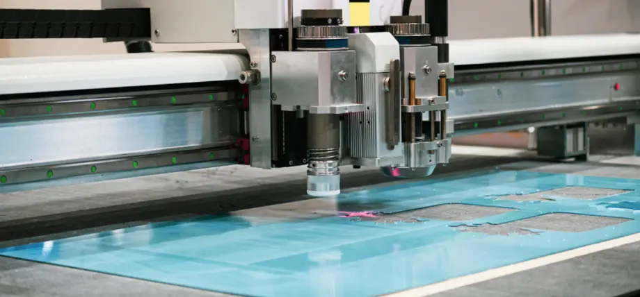 Precision CNC router head with spindle actively milling intricate shapes into a turquoise glass sheet on a flatbed machine.