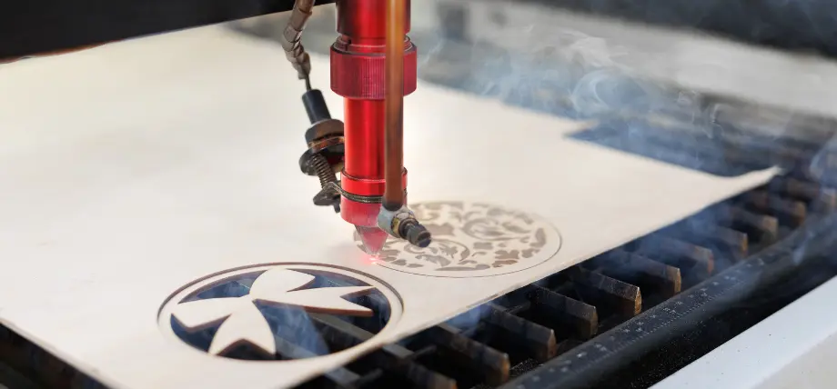 CO2 laser cutter with red head actively engraving and cutting a decorative bow ornament into light plywood, producing visible smoke during laser cutting materials process.