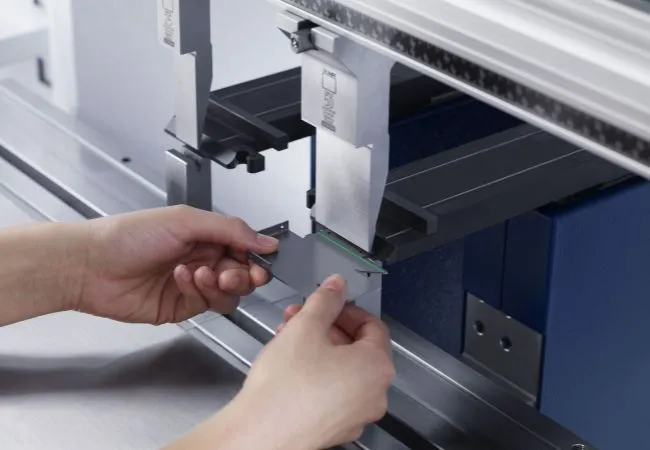 Hands holding a piece of metal being worked on by a machine in a workspace.