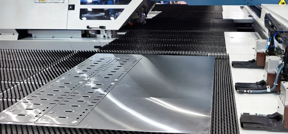 Stainless steel sheet with punched slots and holes positioned on the brush bed of a CNC turret punch press during fabrication.