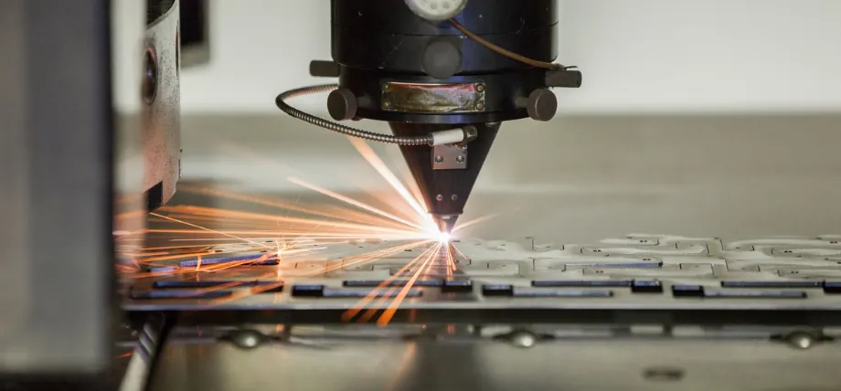 Fiber laser cutting head emitting a bright beam while precisely cutting intricate patterns in thin sheet metal with visible sparks.