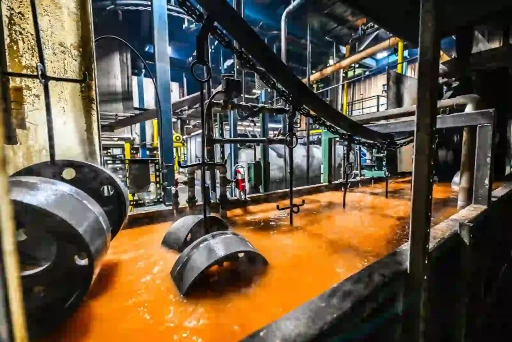 At an industrial facility, metal pieces are placed in an orange electric coating bath, on a conveyor belt.