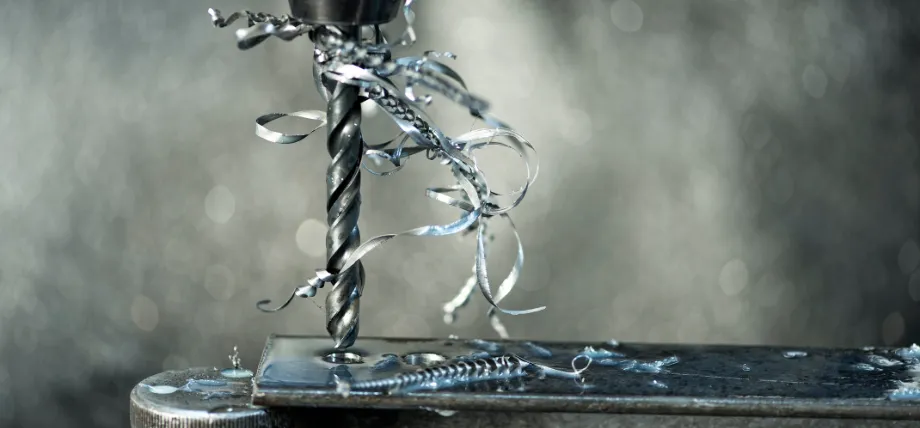 Close-up of a drill bit actively machining a steel workpiece, producing long, shiny metal chips and curls that spiral around the tool in an industrial setting.