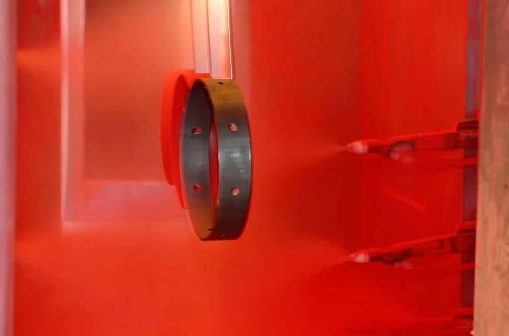 The image shows dry powder coating being applied to metal parts inside an enclosed chamber.