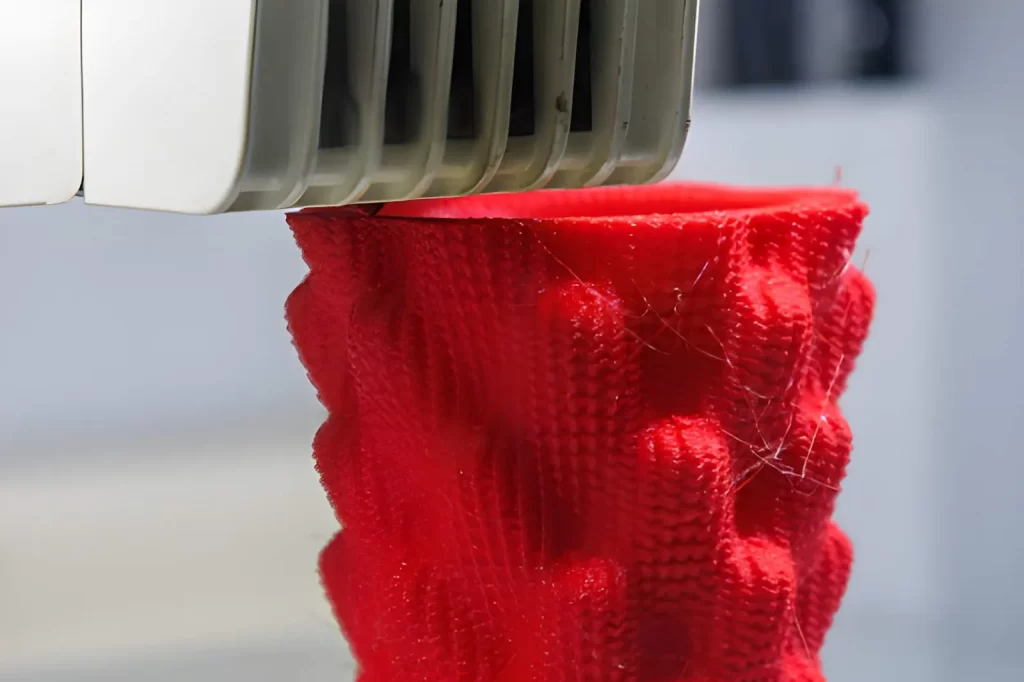 The image shows a close-up of an FDM 3D printer in operation, using additive manufacturing. Thermoplastic material is being deposited layer by layer during a rapid prototyping process to form a precise component.