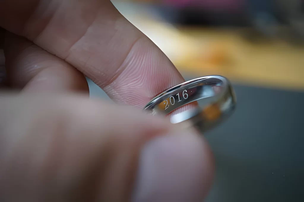 The image shows a male hand holding a stainless steel wedding ring engraved with the year “2016.”
