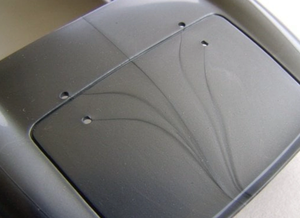 Close-up of a black injection-molded plastic part showing prominent weld lines and flow marks radiating from pin holes, illustrating the aesthetic defects caused by weld lines