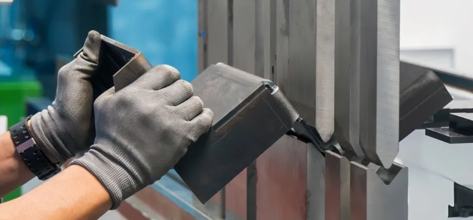 Worker in protective gloves positioning a sheet metal component into a press brake die during sheet metal box fabrication bending process.