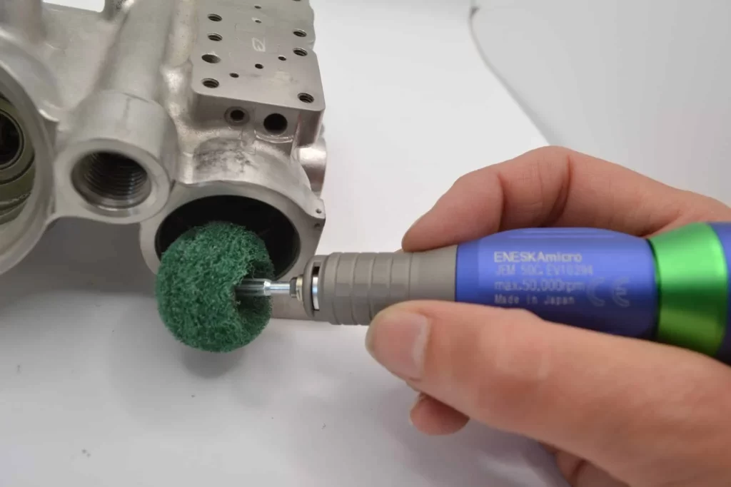 A hand holding a high-speed rotary tool with a green abrasive ball attachment, being used to smooth the inside of a metal engine part