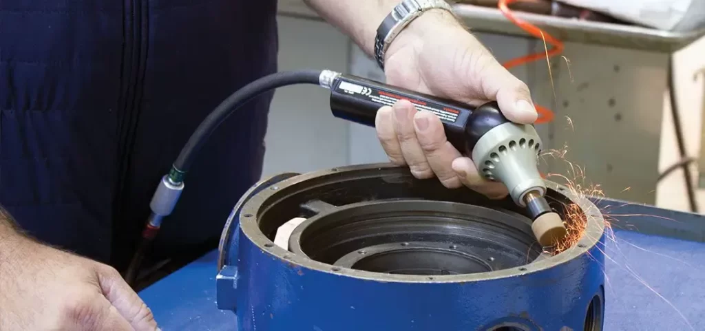 A person using a handheld pneumatic grinding tool to deburr the edge of a large round metal part, with sparks flying