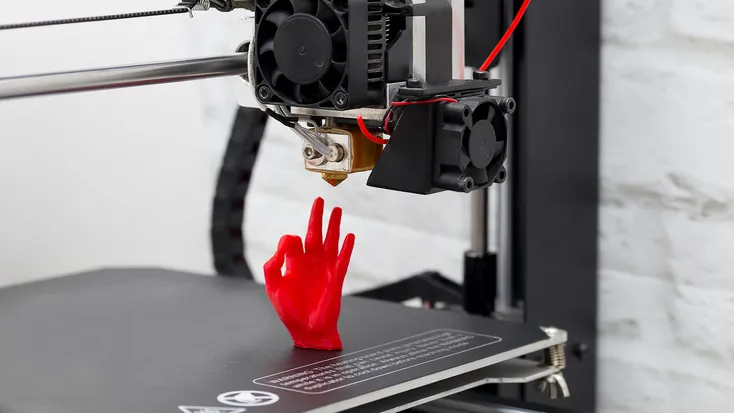 A 3D printer printing a red hand model, with the print head and extruder nozzle visible above the partially completed object on the print bed.