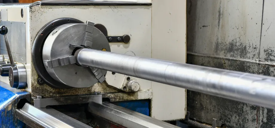 A large cylindrical metal rod is secured in the three-jaw chuck of a heavy-duty industrial lathe machine.
