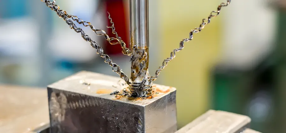 A close-up of a metal drill bit cutting into a steel block, producing long, spiral metallic shavings covered in cutting fluid.