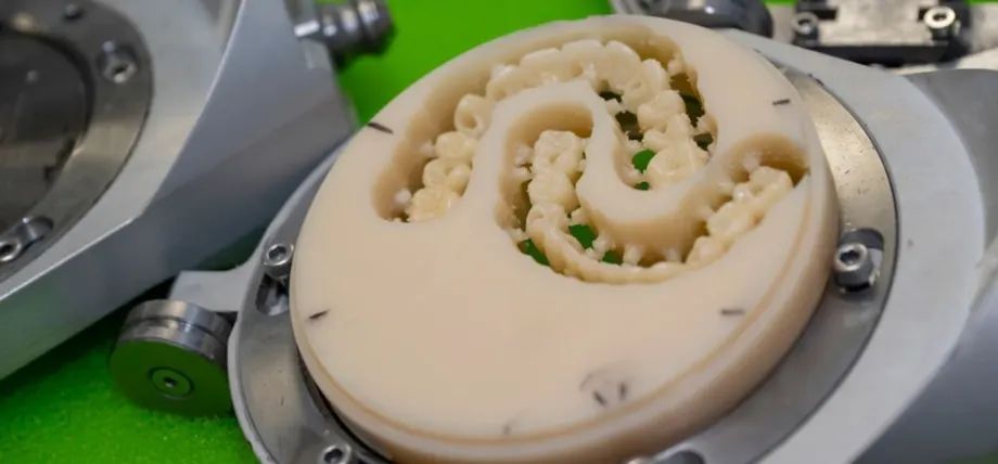 CNC machine milling a beige dental prosthesis with intricate tooth structures on a circular fixture against a green background.