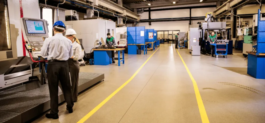 Two professionals in hard hats discussing operations near CNC machines in a clean, organized manufacturing facility with marked walkways and active workstations.