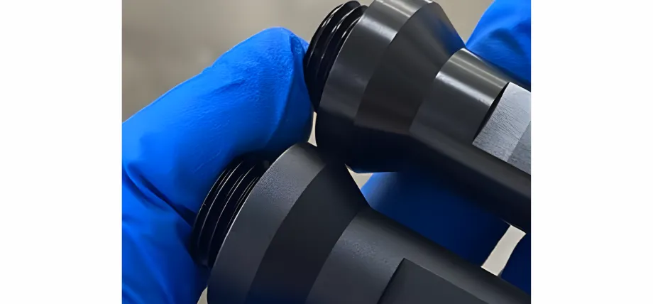 A macro shot held by blue-gloved hands showing the fine matte texture and sharp threads of black anodized aluminum components.