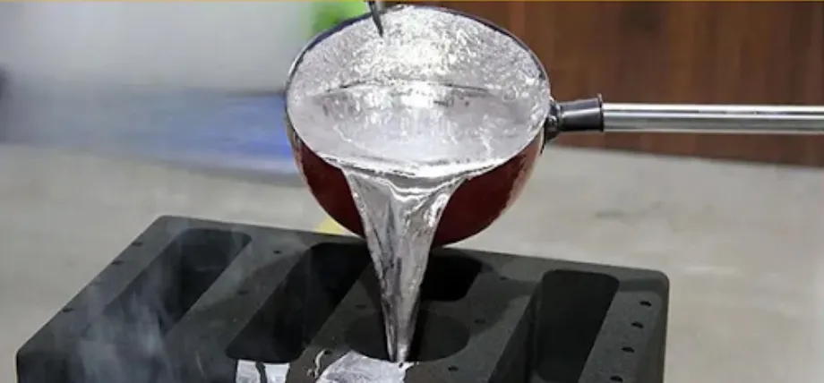 Molten silver-colored metal being poured from a handheld crucible into a dark sand mold.