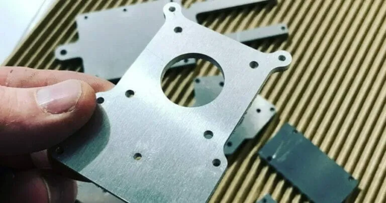A hand holding a laser-cut aluminium mount with holes on the corners and a cutout in the center.