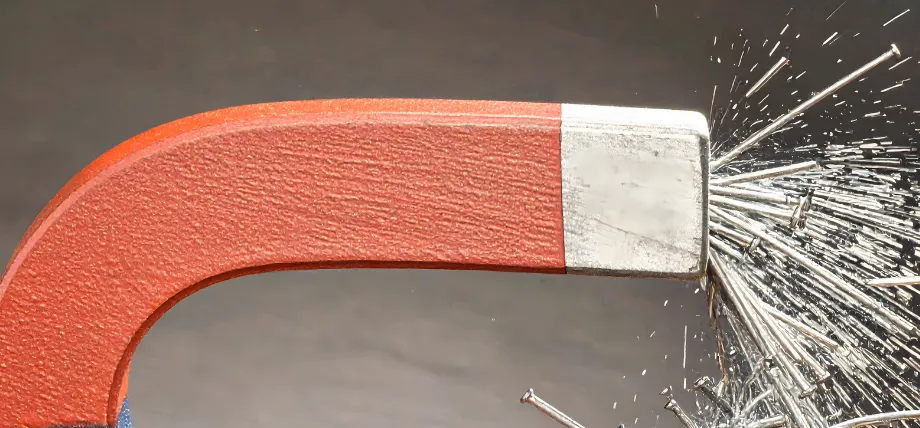 A red horseshoe magnet attracting a cluster of steel nails, demonstrating the magnetic properties of ferrous metals.