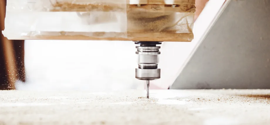 CNC end mill plunging into light wood, producing fine sawdust spray while secured under transparent hold-down tape for clean, precise routing.