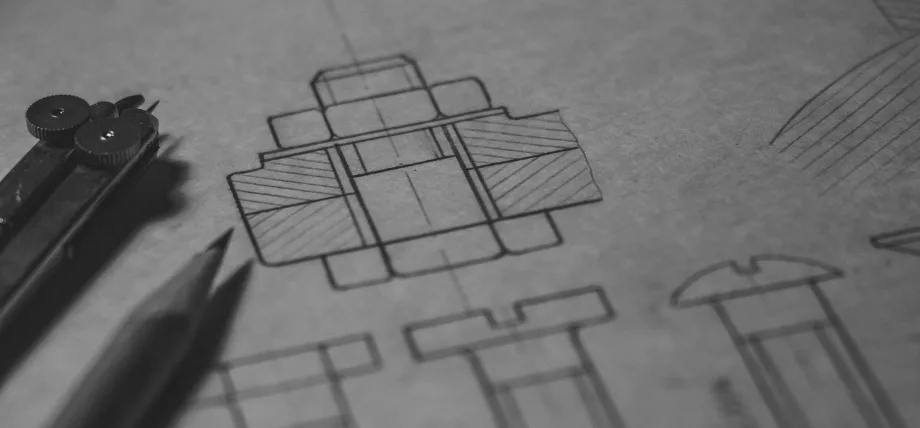 Close-up of mechanical drafting on sepia paper showing orthographic views of a machined component, with compass and pencil in the foreground.